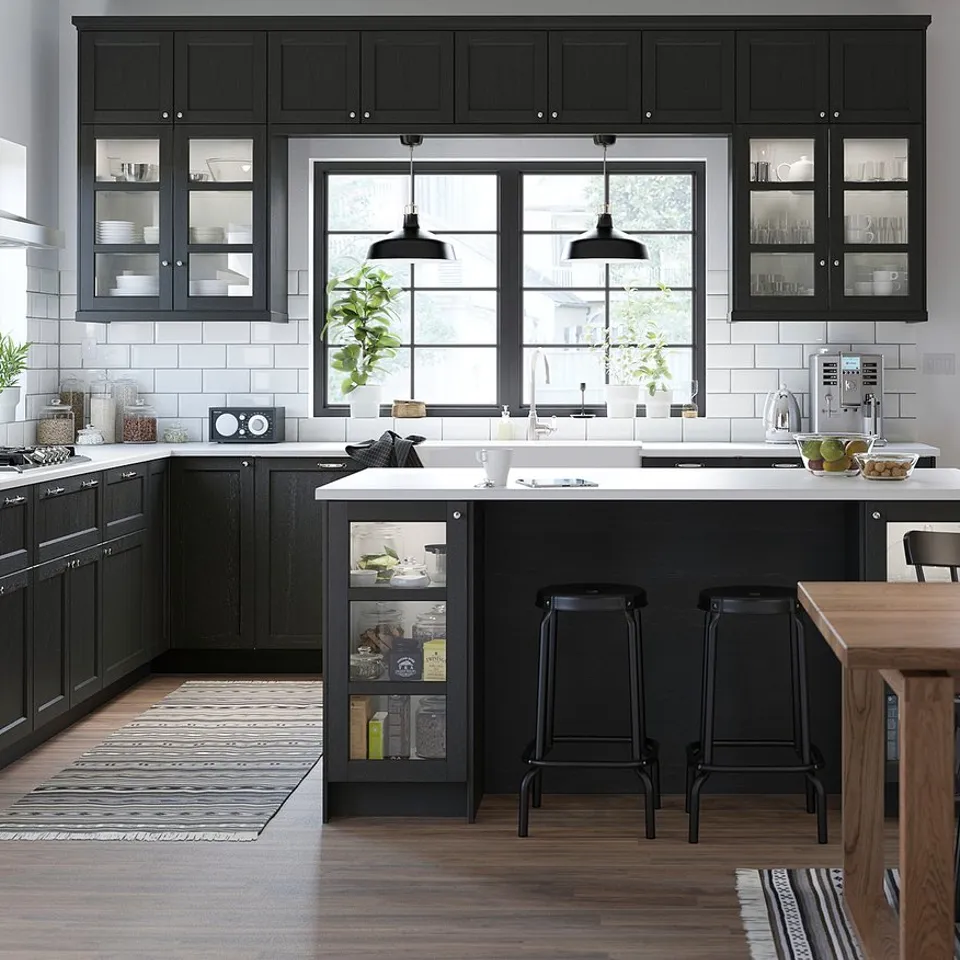 Modern kitchen with black cabinets, white counters, glass cabinets, island, black stools, large windows, pendant lights, black & white rug, plants.