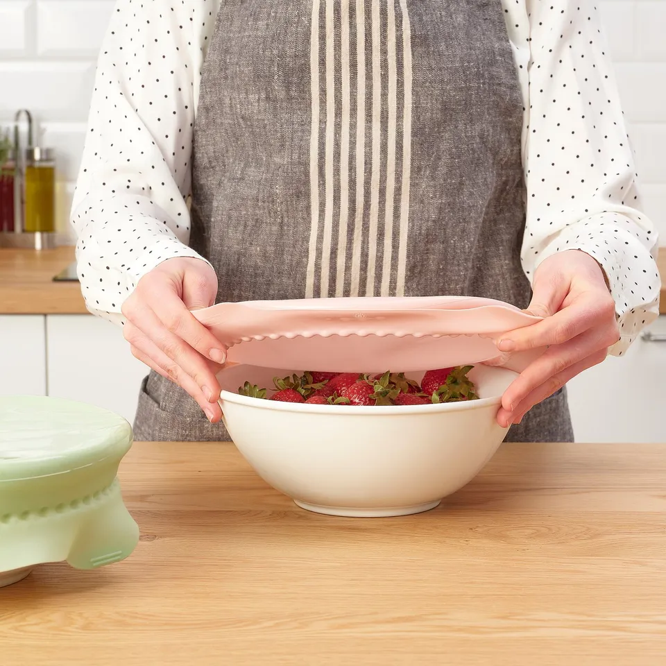 A person placing a light pink bowl cover on a white bowl of strawberries on a wooden table.