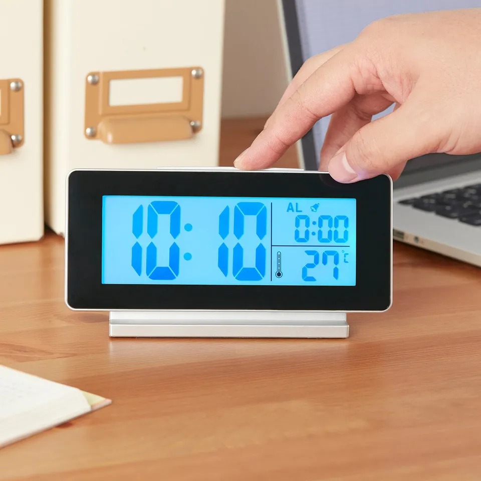 A hand interacts with a modern FILMIS led clock on a wooden desk. The digital display shows time, alarm, and temperature.