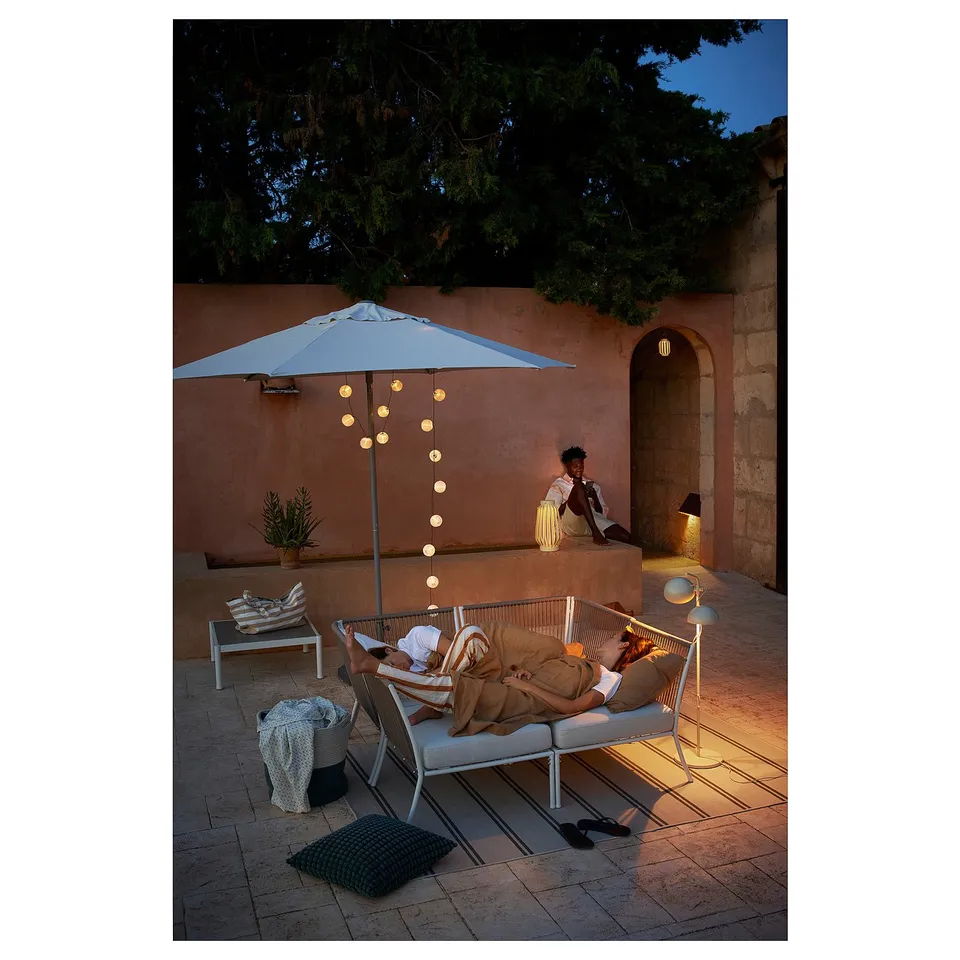 Relaxed patio scene, couple lounging on beige SEGERÖN outdoor sofas under a blue umbrella with string lights, beige and tan cushions, plants, lamp.