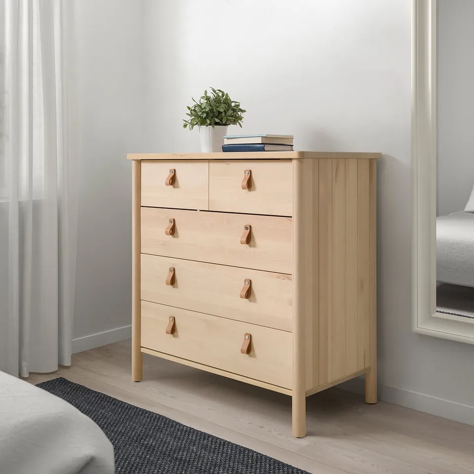 Wooden dresser with 5 drawers, plant, books. Grey wall, bedroom. Anchored.