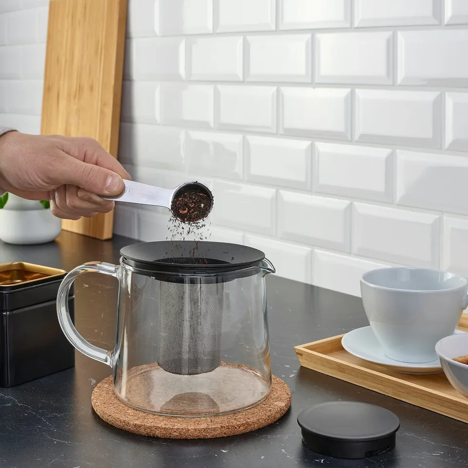 Hand pours tea leaves into RIKLIG glass teapot with mesh infuser on counter.