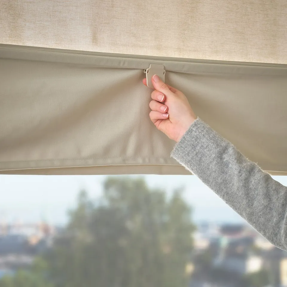 Person adjusting beige RINGBLOMMA roller blind near window, controlling light and privacy.