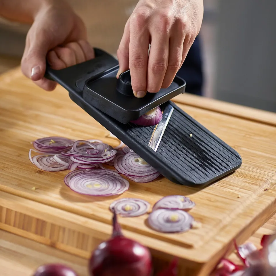 A person slices onions using an IKEA 365+ mandoline slicer on a wooden board.
