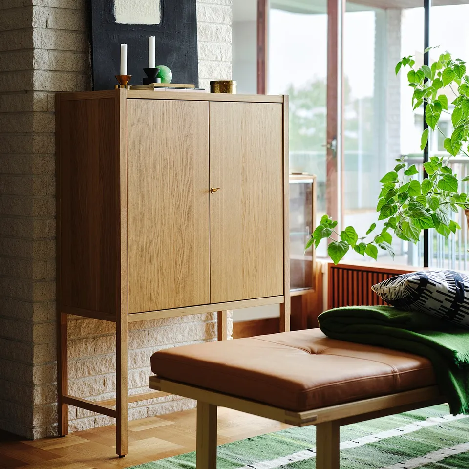 Light wood STOCKHOLM 2025 cabinet, brick wall, bench, green pillow, plant.