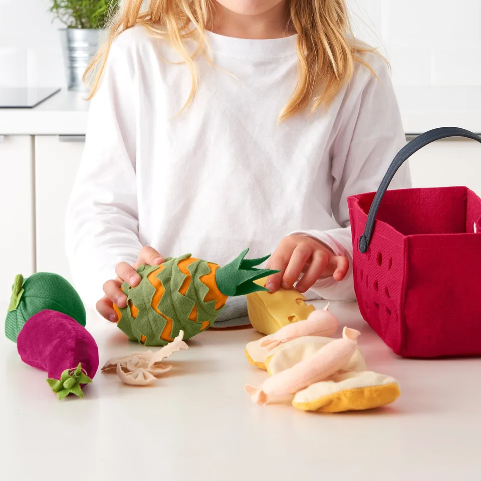 Child holds fabric pineapple with play food set. Includes beets, fish, cheese, sausages, bread, & shopping basket.
