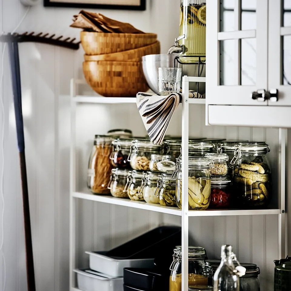 Glass jars filled with pickles and jams on shelves, with a glass serving as a beverage dispenser in a white kitchen.