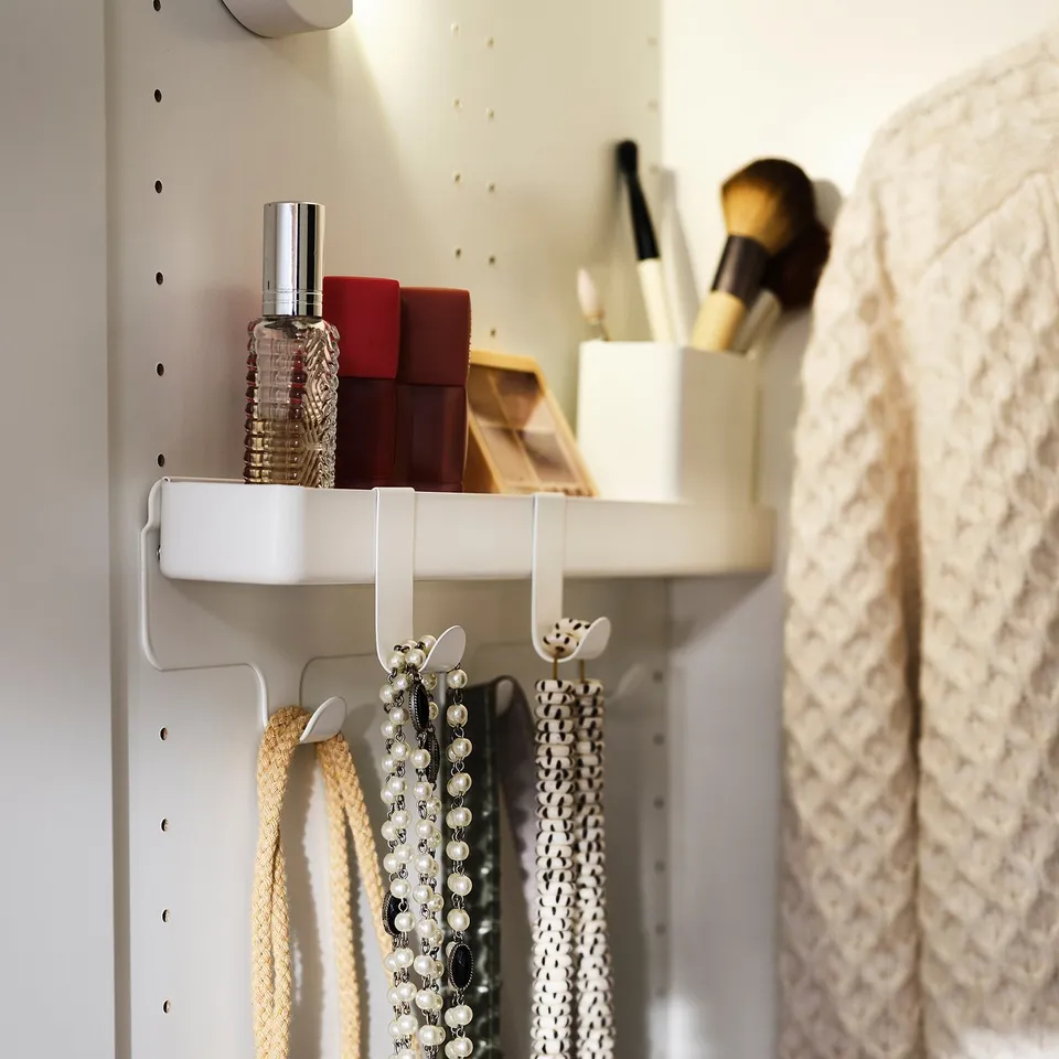 A white wall-mounted storage unit with hooks holding necklaces and a shelf displaying beauty products.
