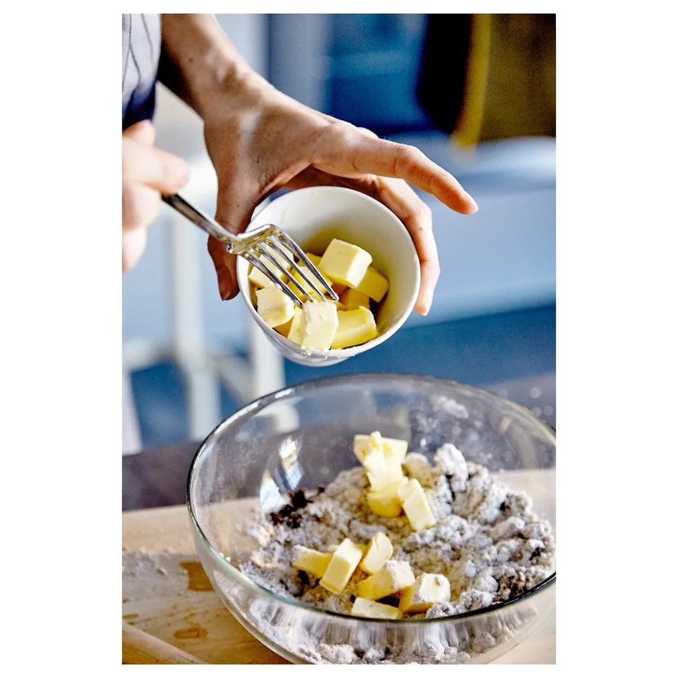 A person uses a pastry cutter to incorporate butter into a mixture in a glass bowl on a blue countertop.