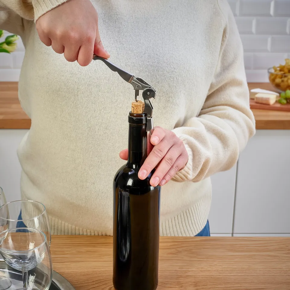 Person using a corkscrew to open a wine bottle on a wooden table.
