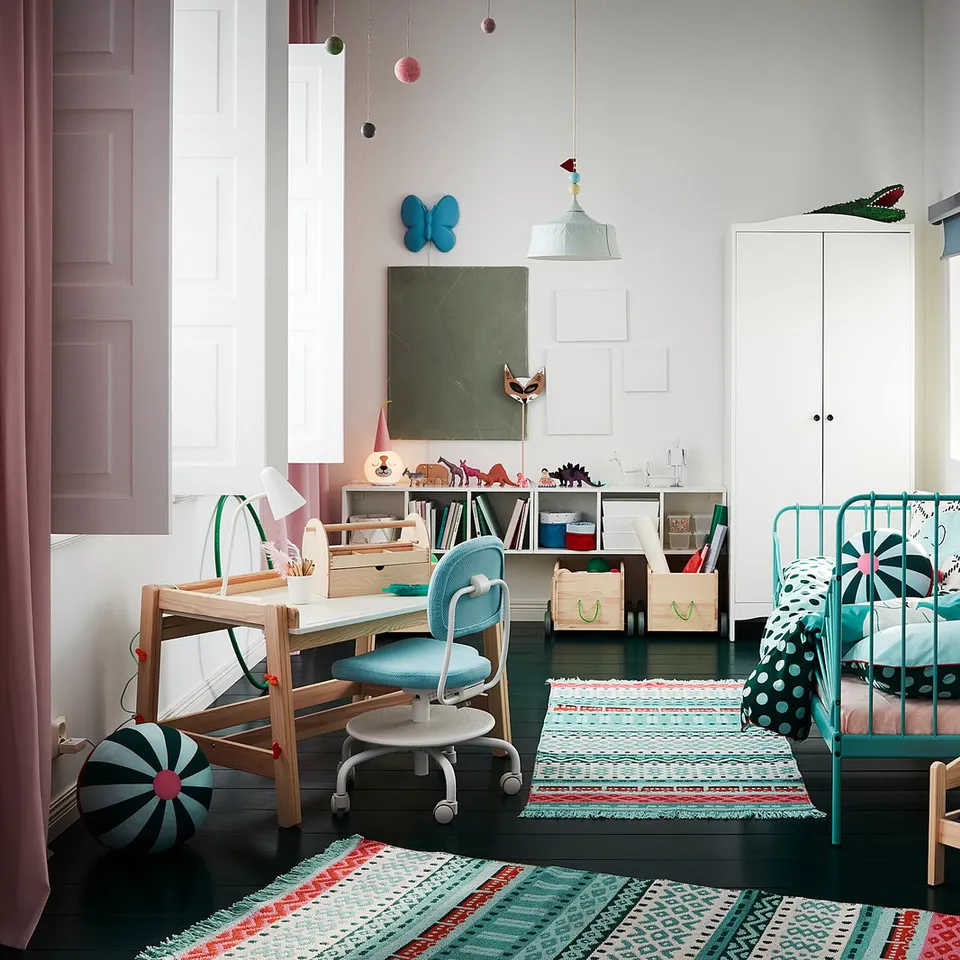 Colourful kids room with a wooden desk, blue chair, green bed, shelves, and rugs. Wall hangings and decorations.