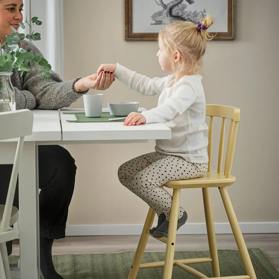 Child on yellow AGAM chair, interacting with adults hand, green bowl and cup on table.