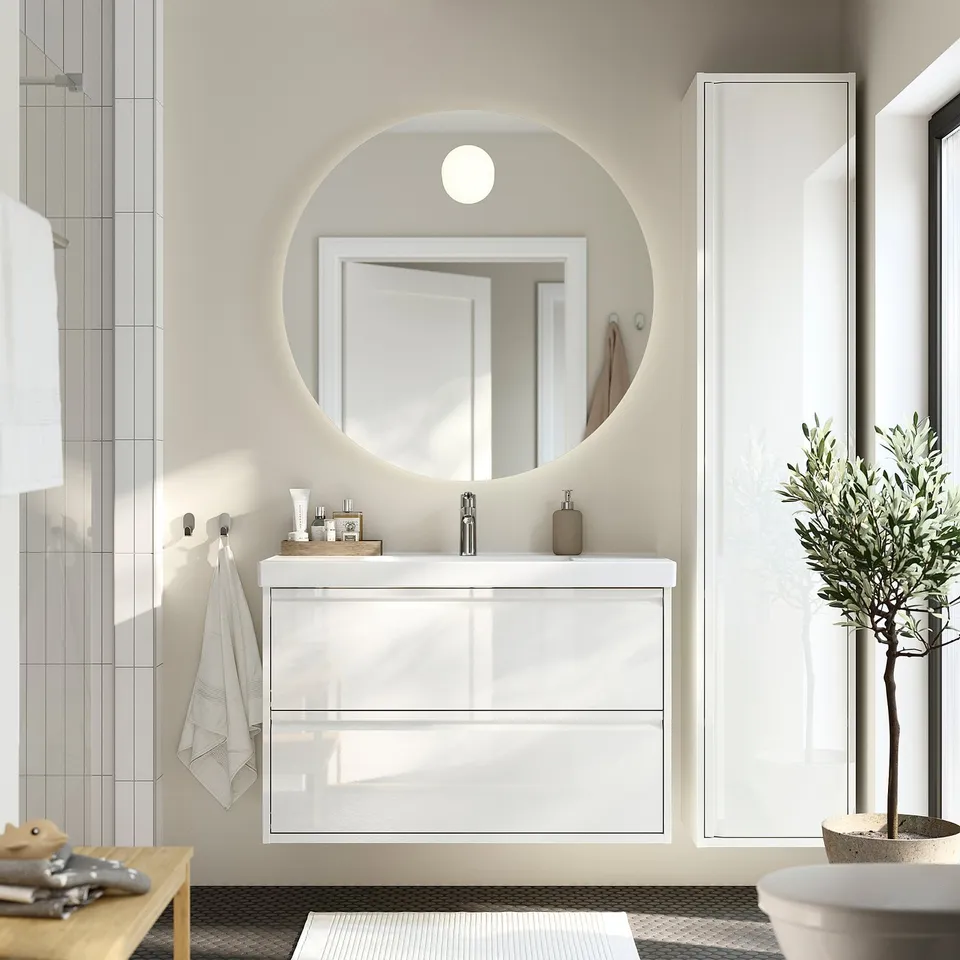 Modern white ÄNGSJÖN bathroom vanity, round mirror, glass doors, sleek handles. Dark floor, green plant.