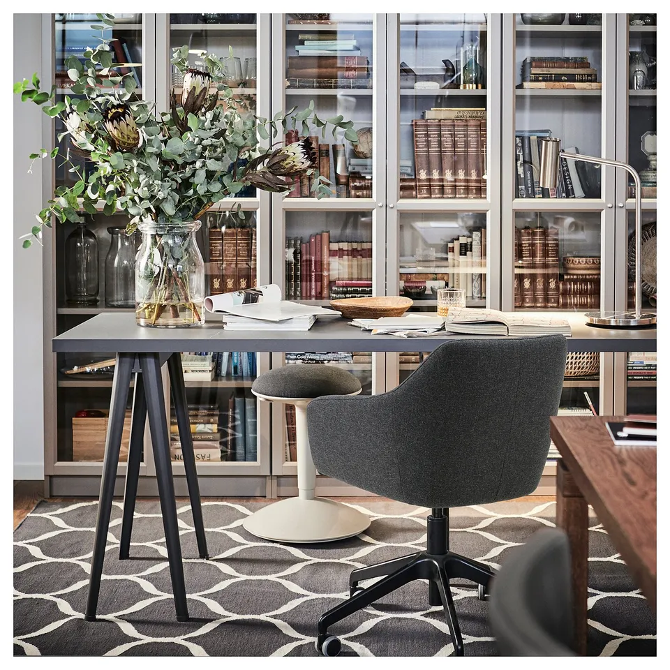 Home office setup with TOSSBERG grey swivel chair, black desk, glass vase with greenery, bookshelves, and geometric rug.