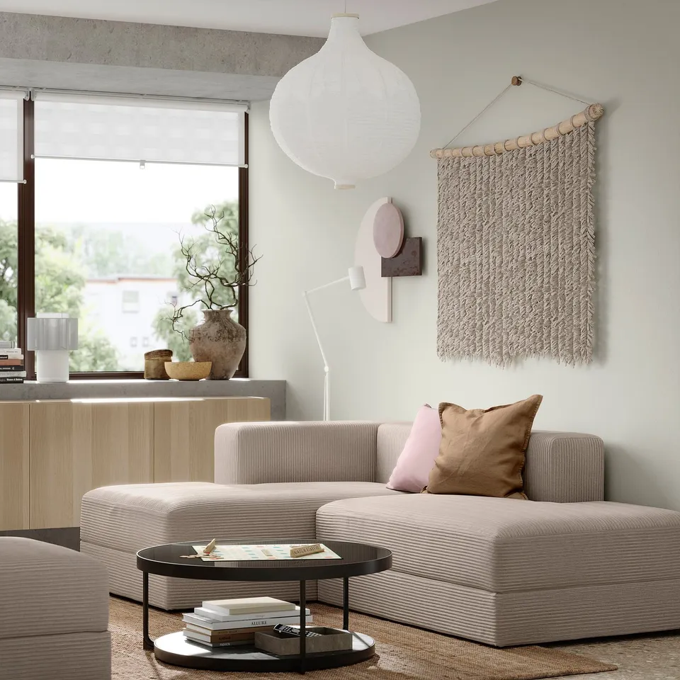 Modern living room with JÄTTEBO beige modular sofa, pink and brown cushions, hanging light, wall art, wooden cabinet, and large window.