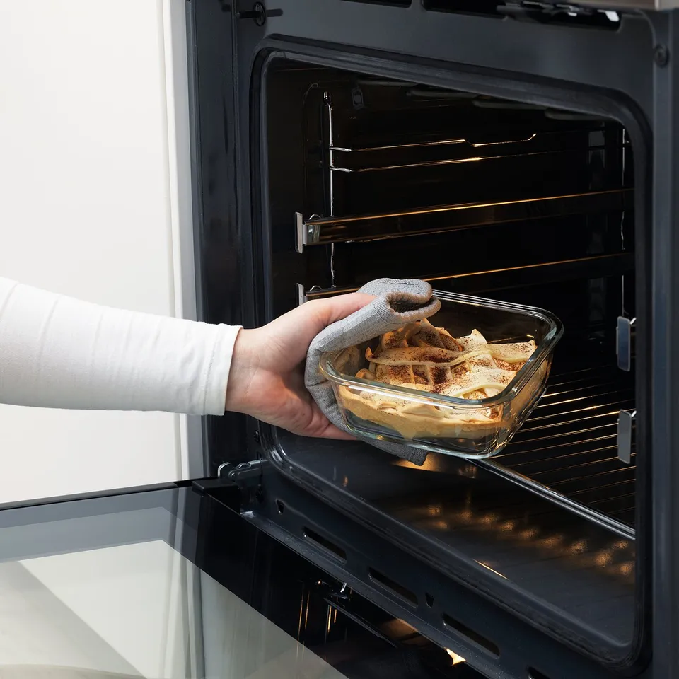 Person wearing oven gloves placing dish into oven. The dish is a transparent food container made from oven-safe glass.