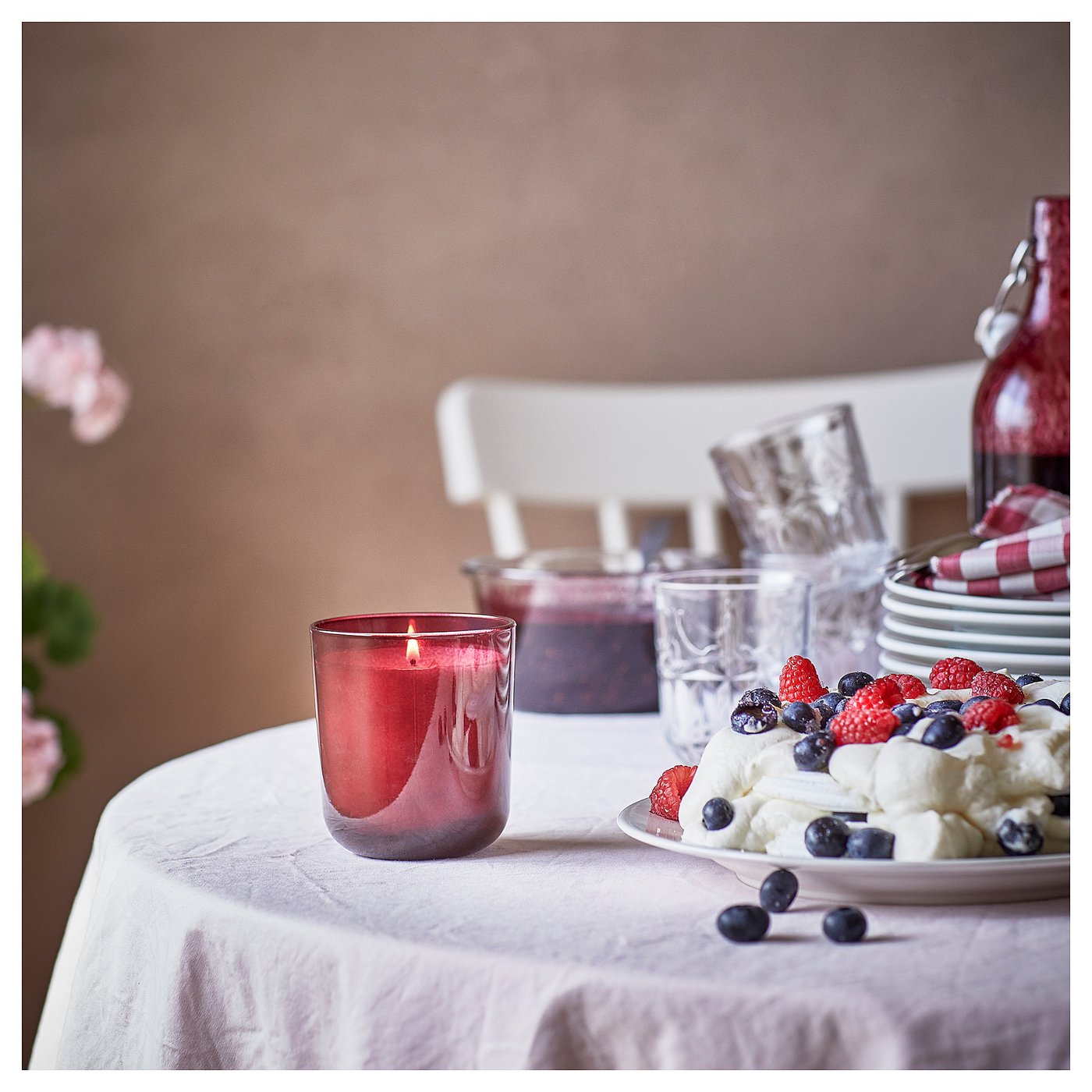 A red glass STÖRTSKÖN candle with lit wick on a table. Table has cream dessert, berries, glasses, and stacked plates. White chair in background.