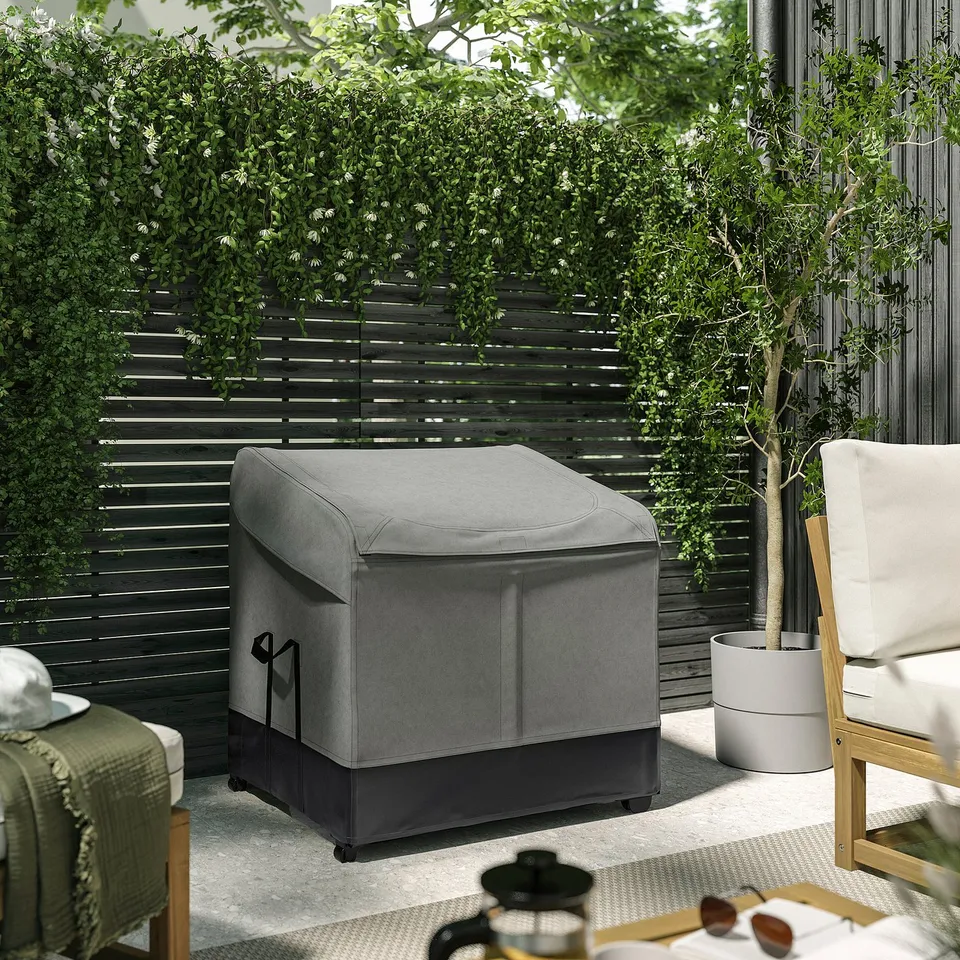 A grey VÄTTERSÖ outdoor storage box on a patio. Its surrounded by lush greenery and furniture, demonstrating its size and outdoor use.