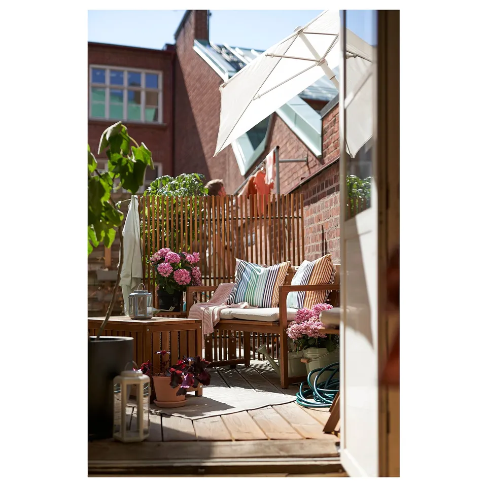 Cosy balcony with wooden NÄMMARÖ furniture, white cushions, pink flowers, and potted plants against a brick backdrop.