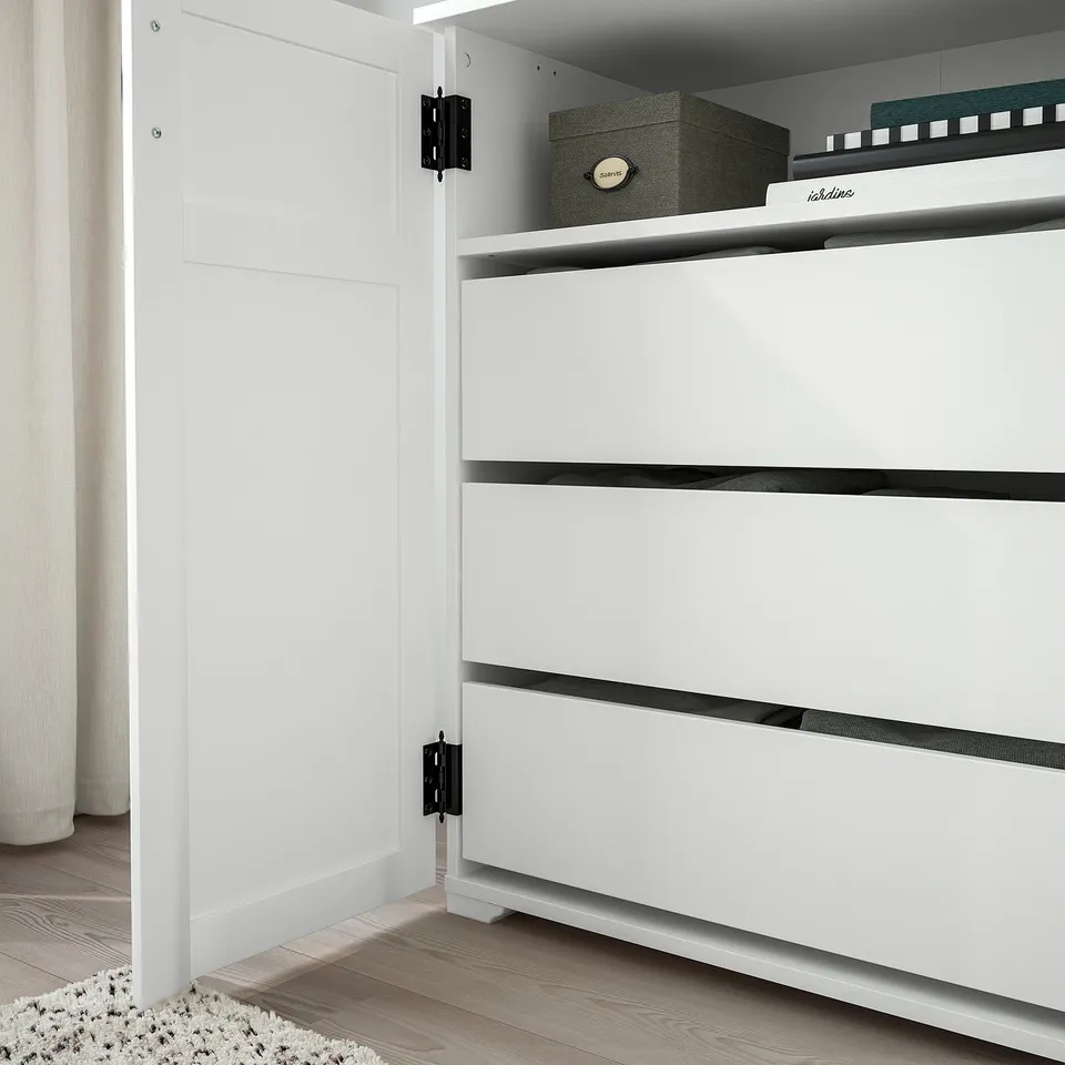 White cabinet with three drawers, black handles, and hinges. One shelf on top holding storage boxes. Floor has rug.