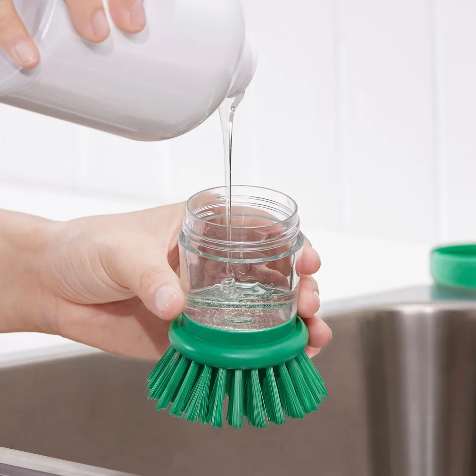 Person pouring washing-up liquid into a jar brush. The brush has a green handle, designed for easy grip and cleaning tough spots.
