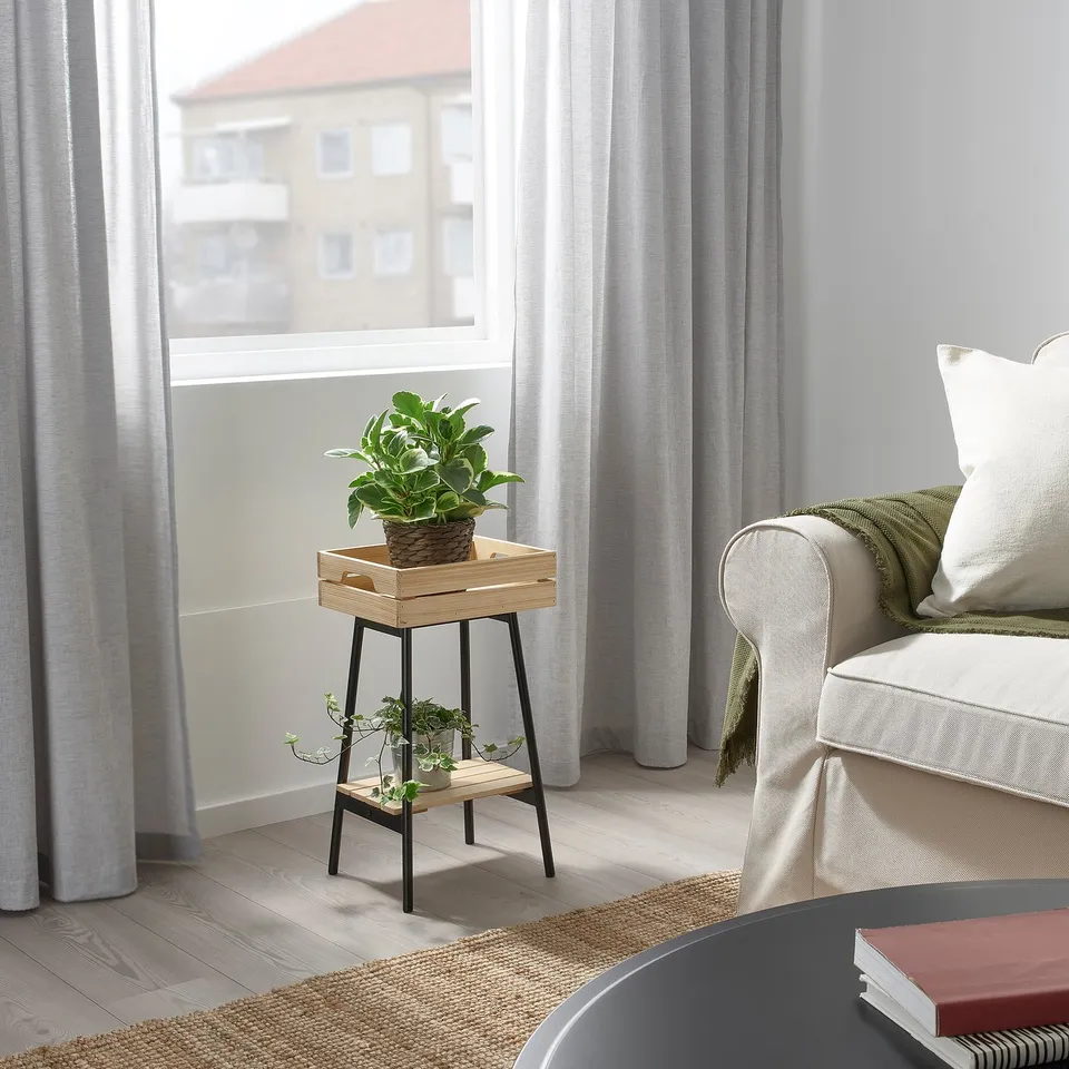 Small black metal & wood plant stand near window, holding green plant. Beside white armchair with blanket & pillow, under large window with curtains.