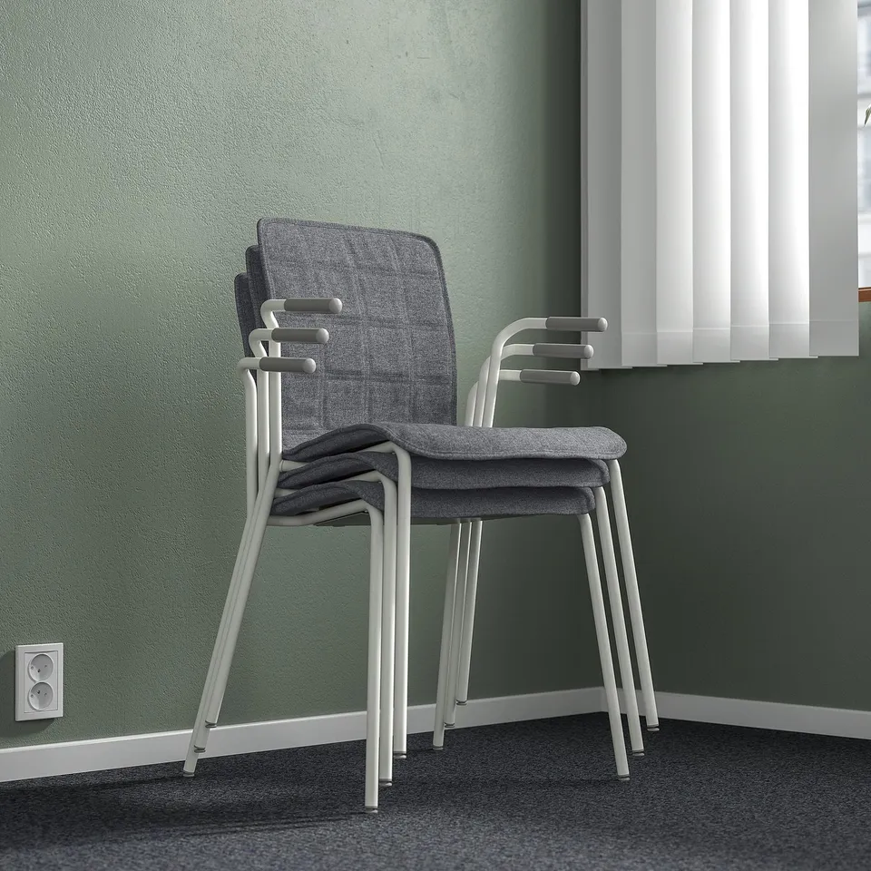 Stacked modern conference chairs with grey fabric and white frames, shown against a green wall near a window with blinds.