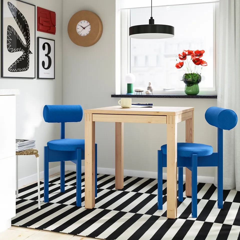 Small blue IKEA PS 2026 chairs & table set. Bright blue fabric seats & wooden frame. Next to a window & a clock.