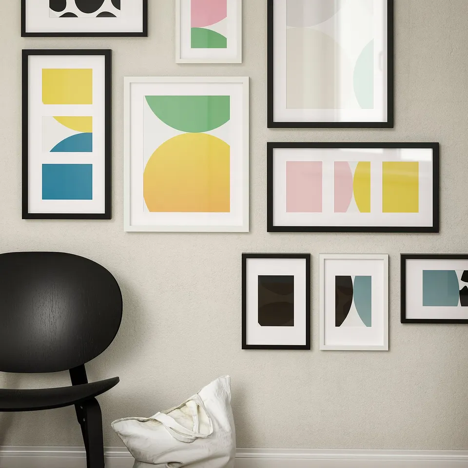 Modern art gallery wall with multiple abstract, colourful frames in black and white, varying sizes. A black chair and white pillow in foreground.