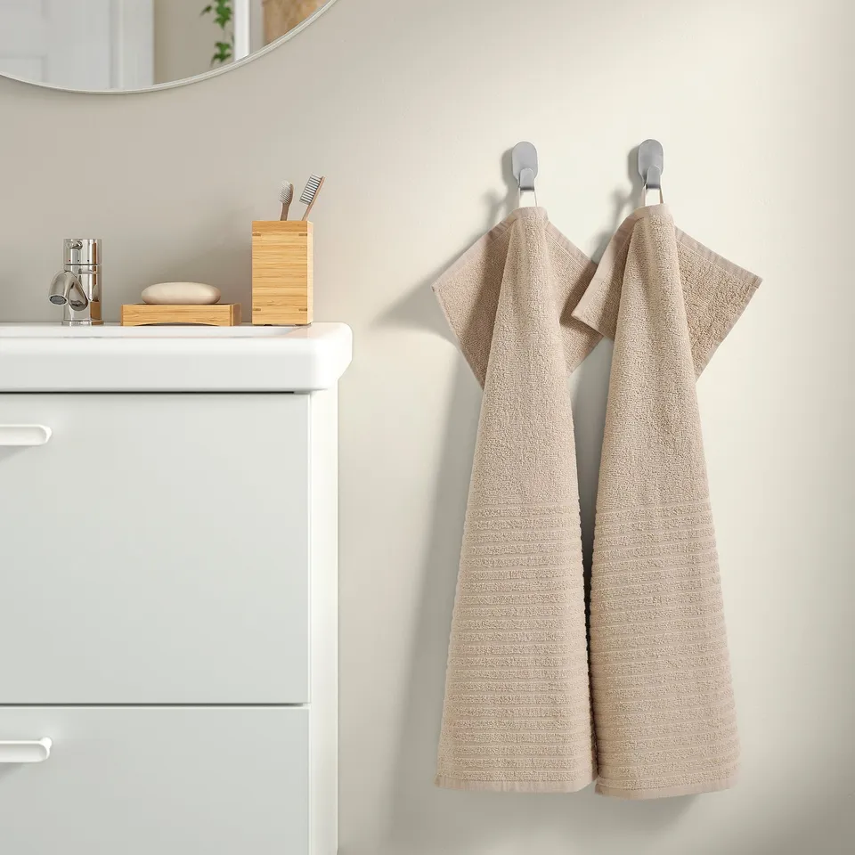 Two beigevägsjön cotton towels hang beside a white sink, with a bamboo toothbrush holder, mirror, and towels on a countertop.