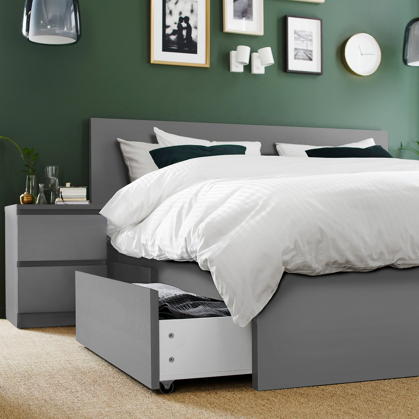 Modern grey bed with white linens, dark headboard, and pull-out storage drawers, set against a green wall with decorations.