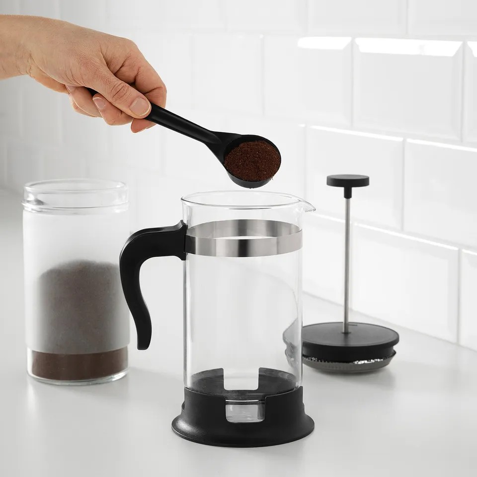 Hand pours coffee grounds into a clear glass UPPHETTA cafetière. The plunger and handle are visible, ready for brewing coffee.