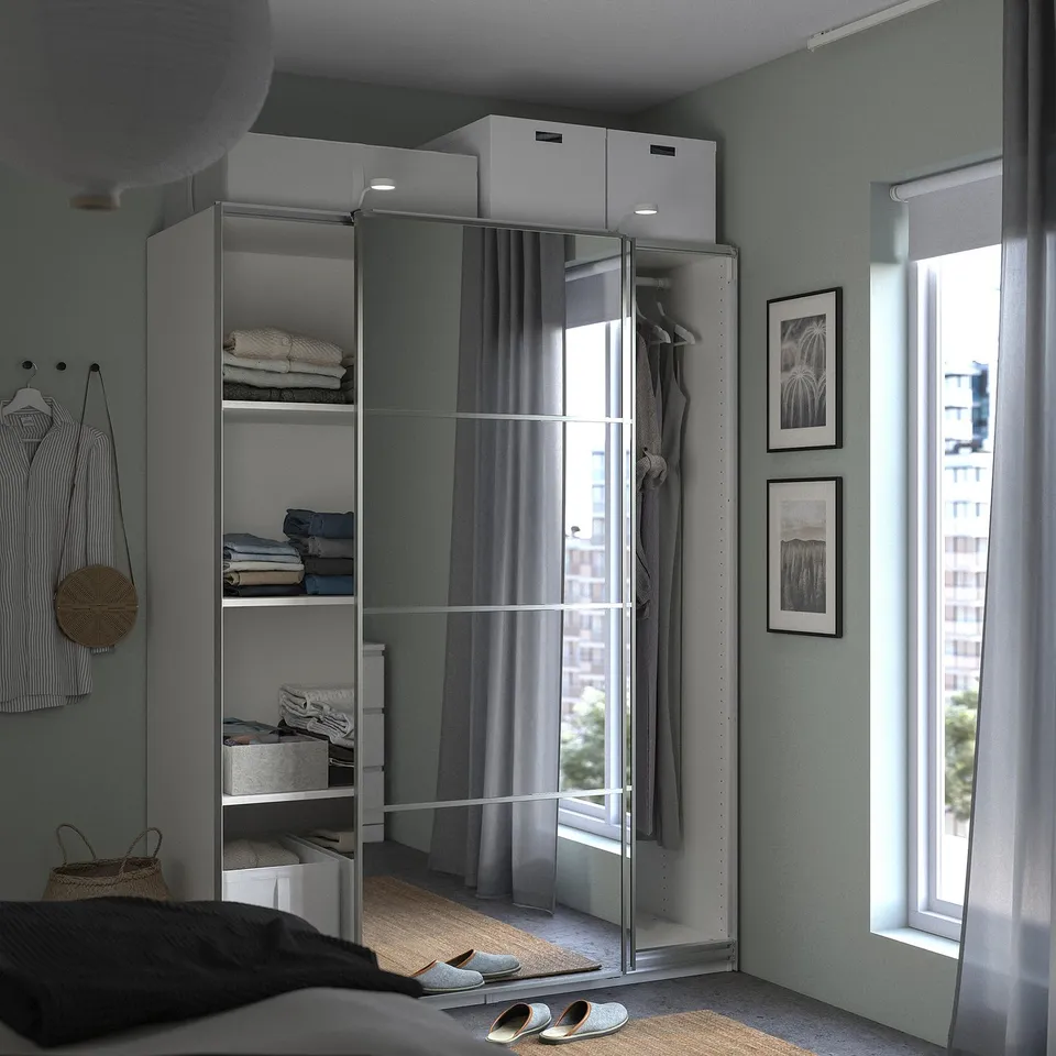 Modern bedroom with PAX wardrobe, mirrored doors, grey curtains, clothes, and art by bright window.