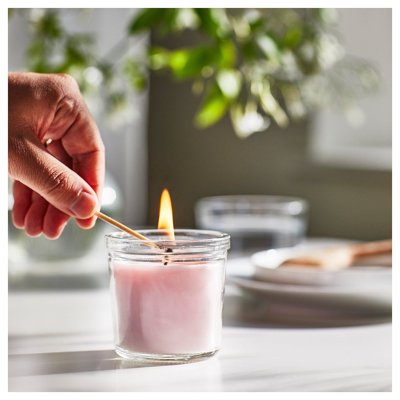 Hand lights pink candle in clear glass jar on white table; green plant background.