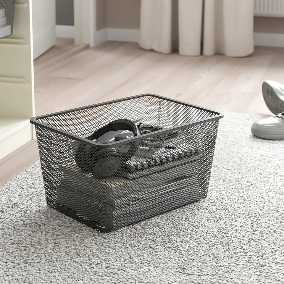 Metal mesh basket, holding headphones on carpeted floor. Organises tech items, part of TROFAST series.