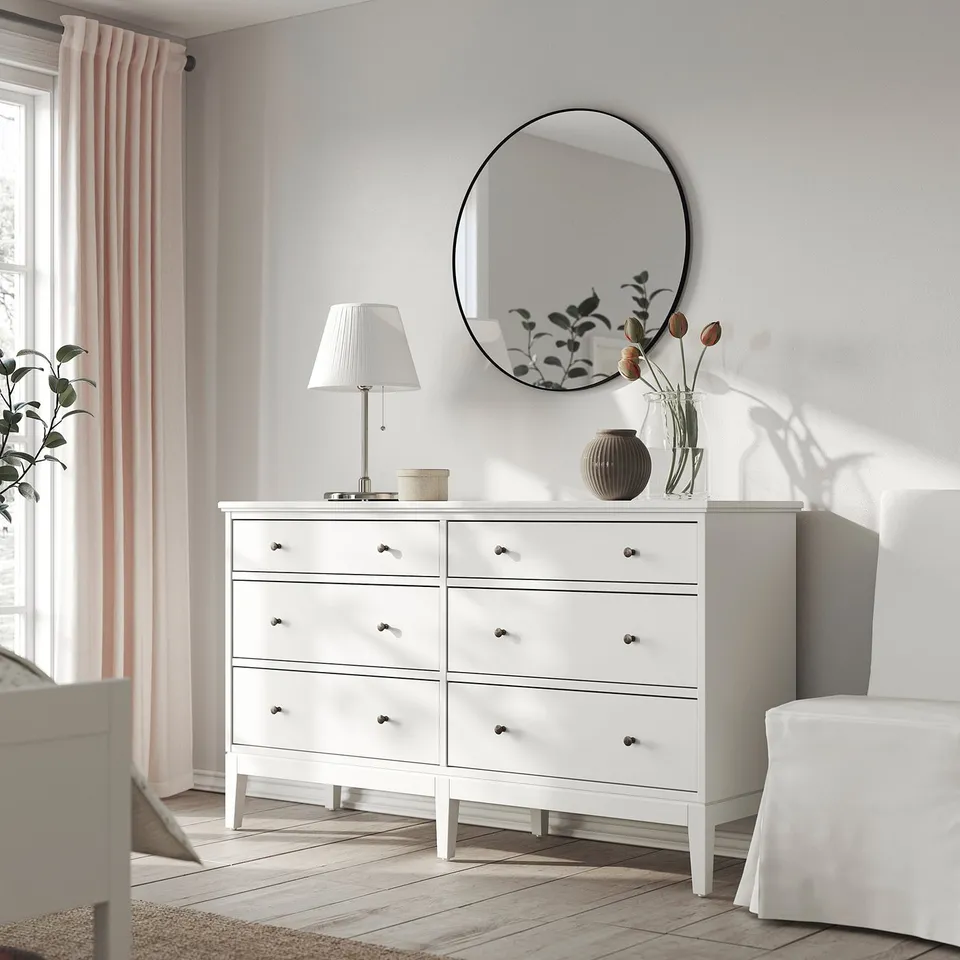 Modern white bedroom with round mirror, white dresser, lamp, and flowers.