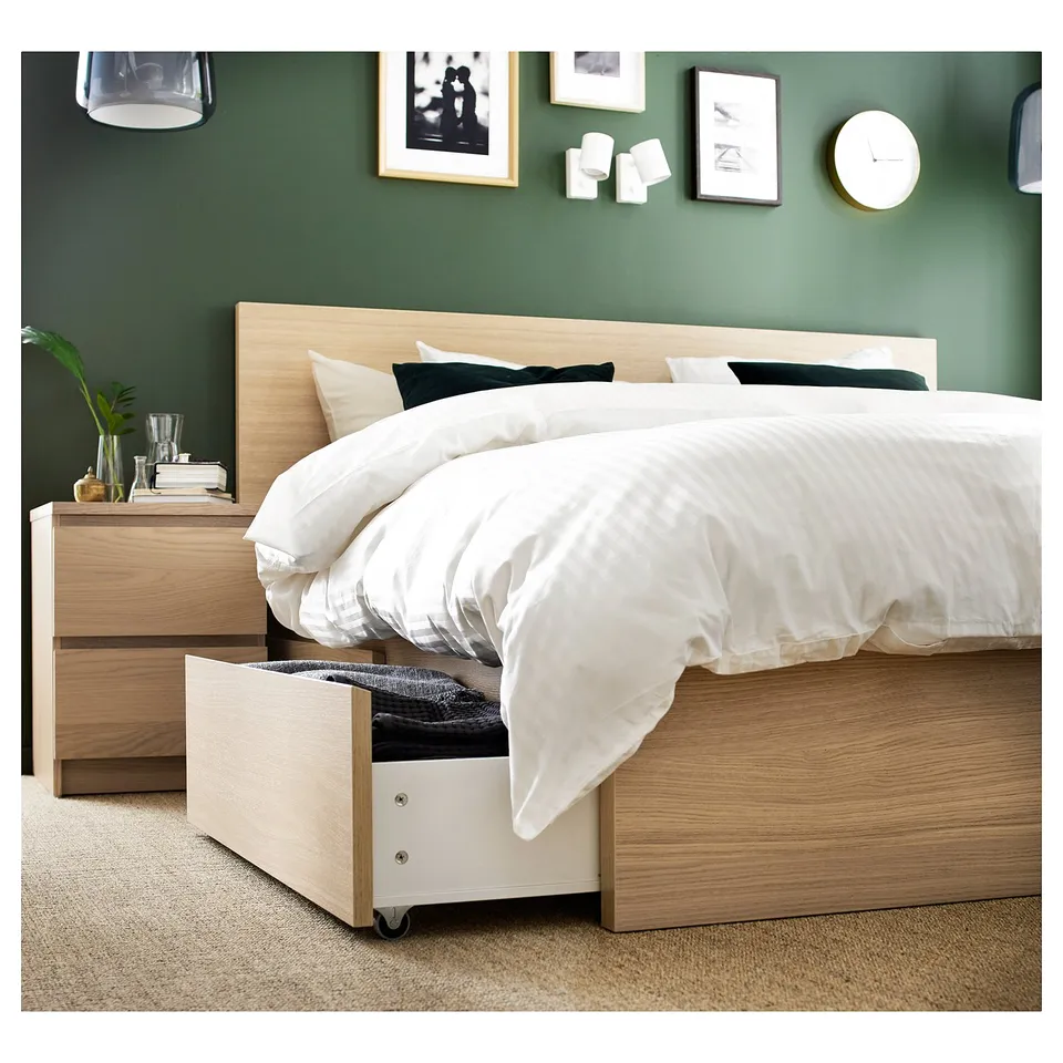 Wooden MALM bed against green wall, white bedding, green pillows, open drawer with black linens, bedside table with plants and books.