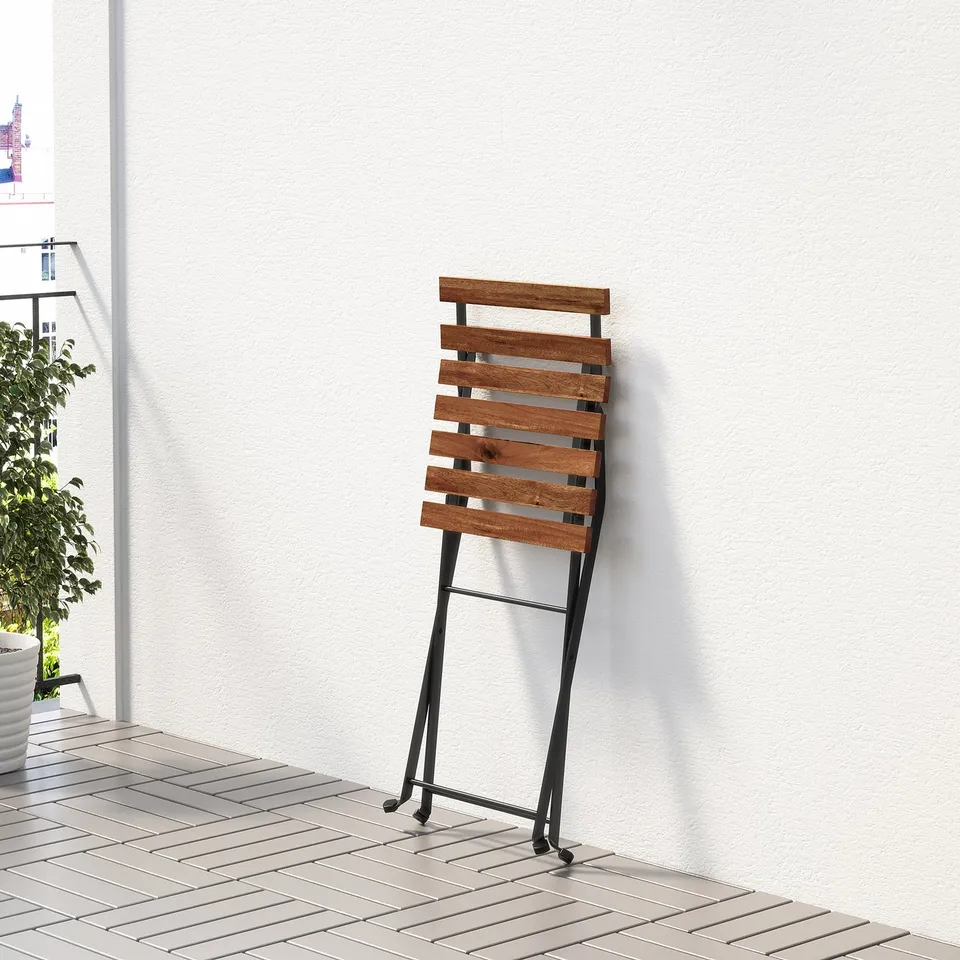 Wooden folding chair leans against a white wall on a tiled patio. Chair is stylish with slatted design.
