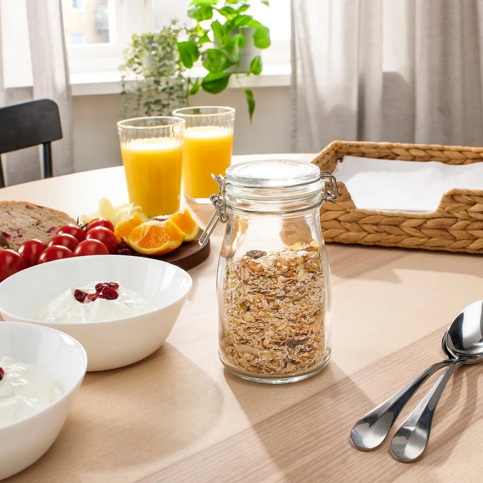 A table set with breakfast items. Jars of granola, bowls of yoghurt with berries, glasses of orange juice, and spoons are arranged neatly.