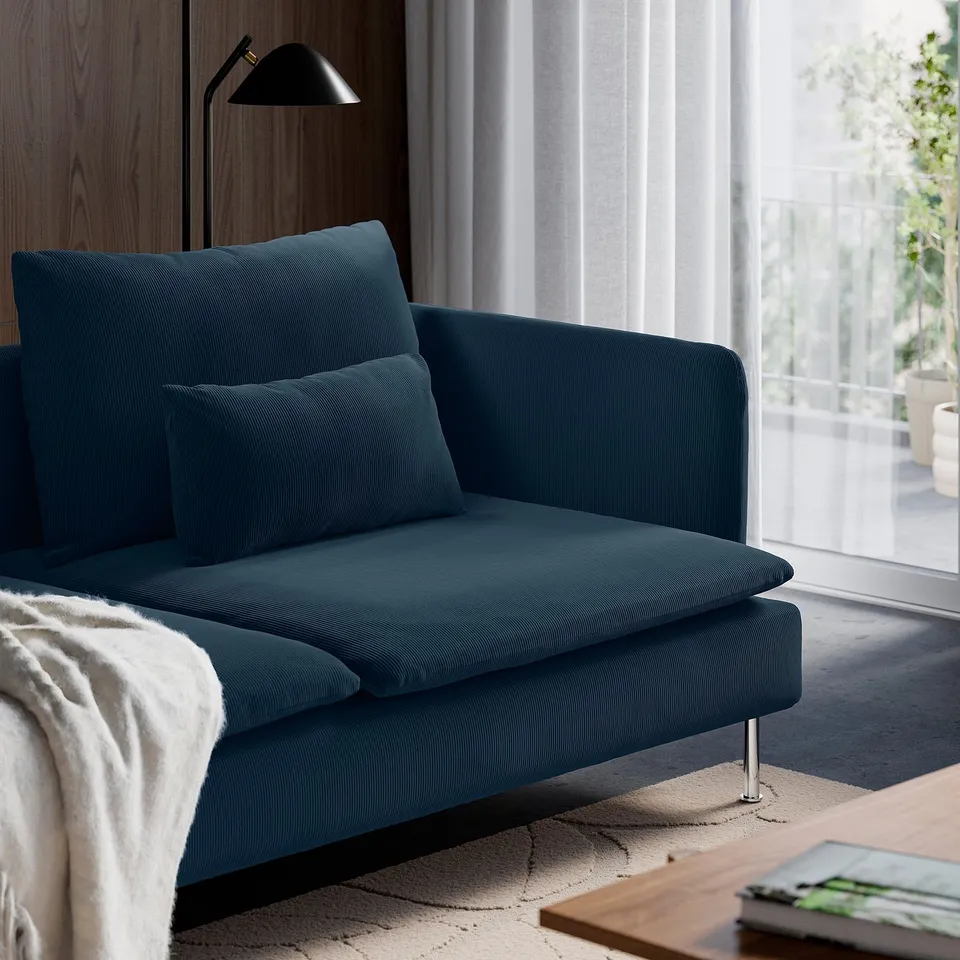 Dark blue SÖDERHAMN sofa with plush cushions, patterned rug, and modern lamp. Studio setting with large window and light curtains.
