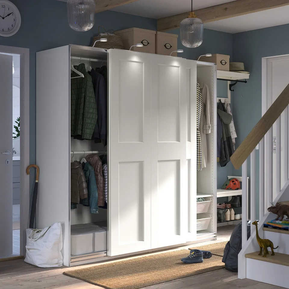 A modern closet named PAX, featuring white sliding doors and built-in lights, neatly storing various clothes and accessories.