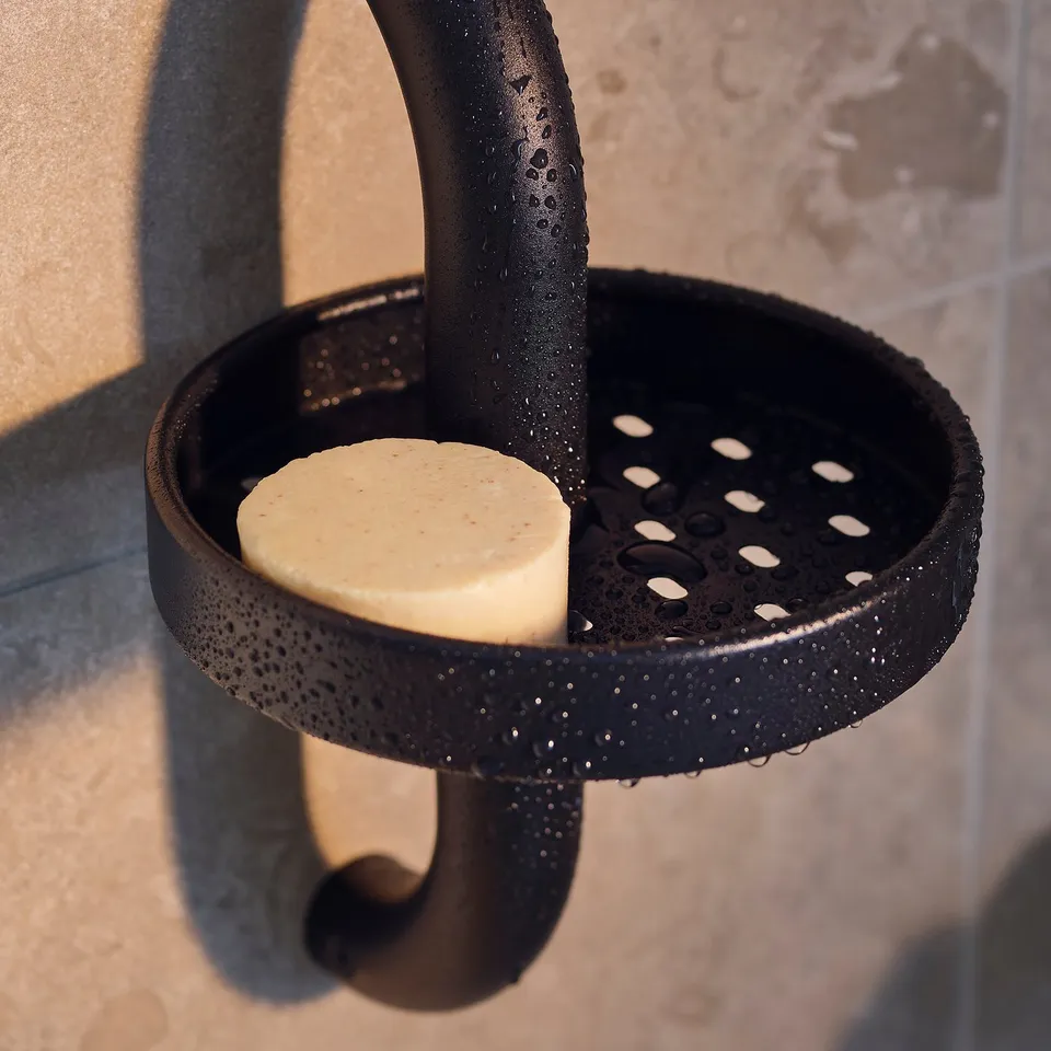 Black BÄSINGEN shower shelf with round soap, grey tiled wall.