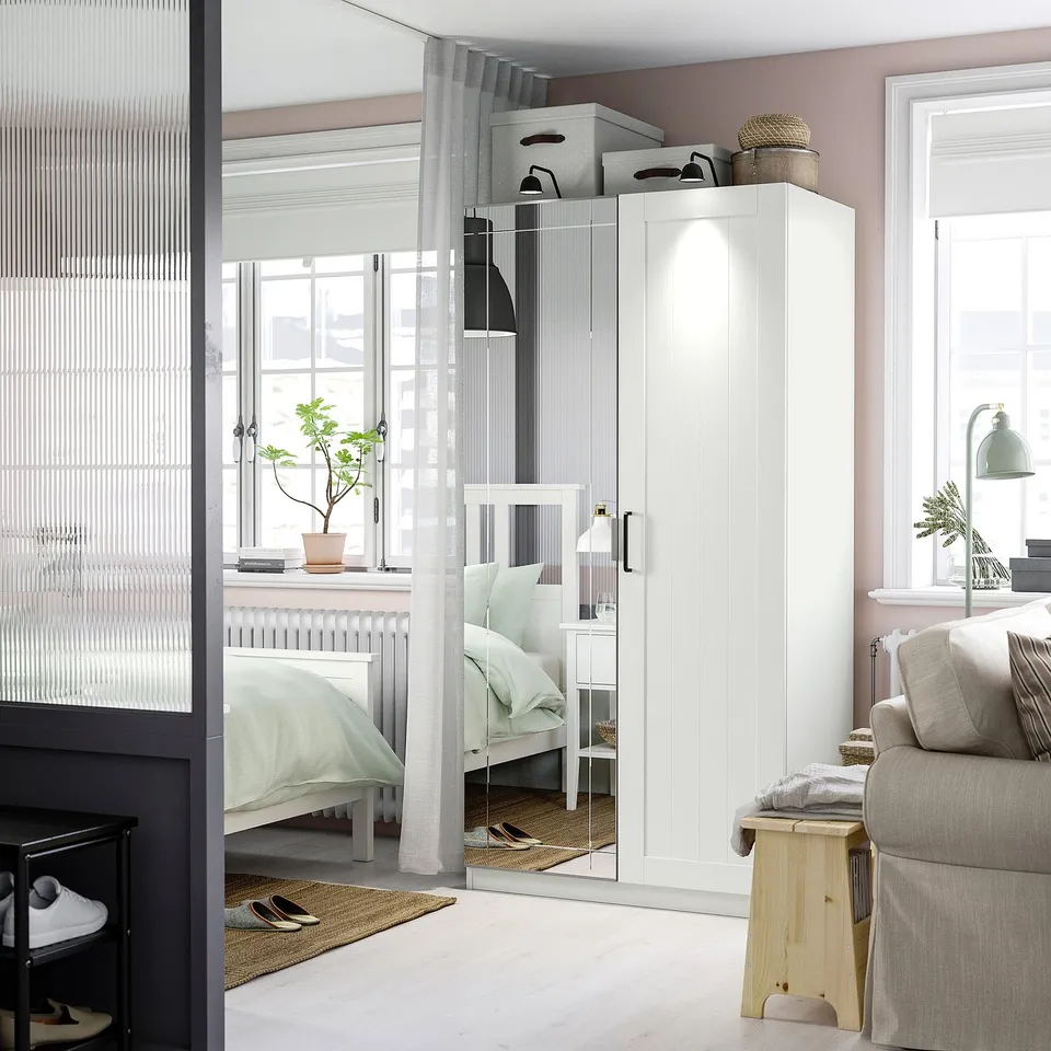 Modern bedroom with white PAX wardrobe, large window, mirrored doors, and cosy decor including storage boxes, a basket, grey sofa, and wooden stool.