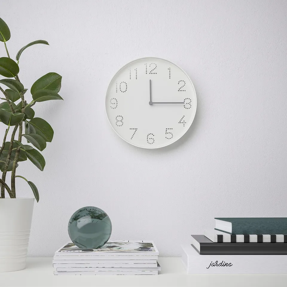 Modern white wall clock at 12:30, above books, green sphere, plant.