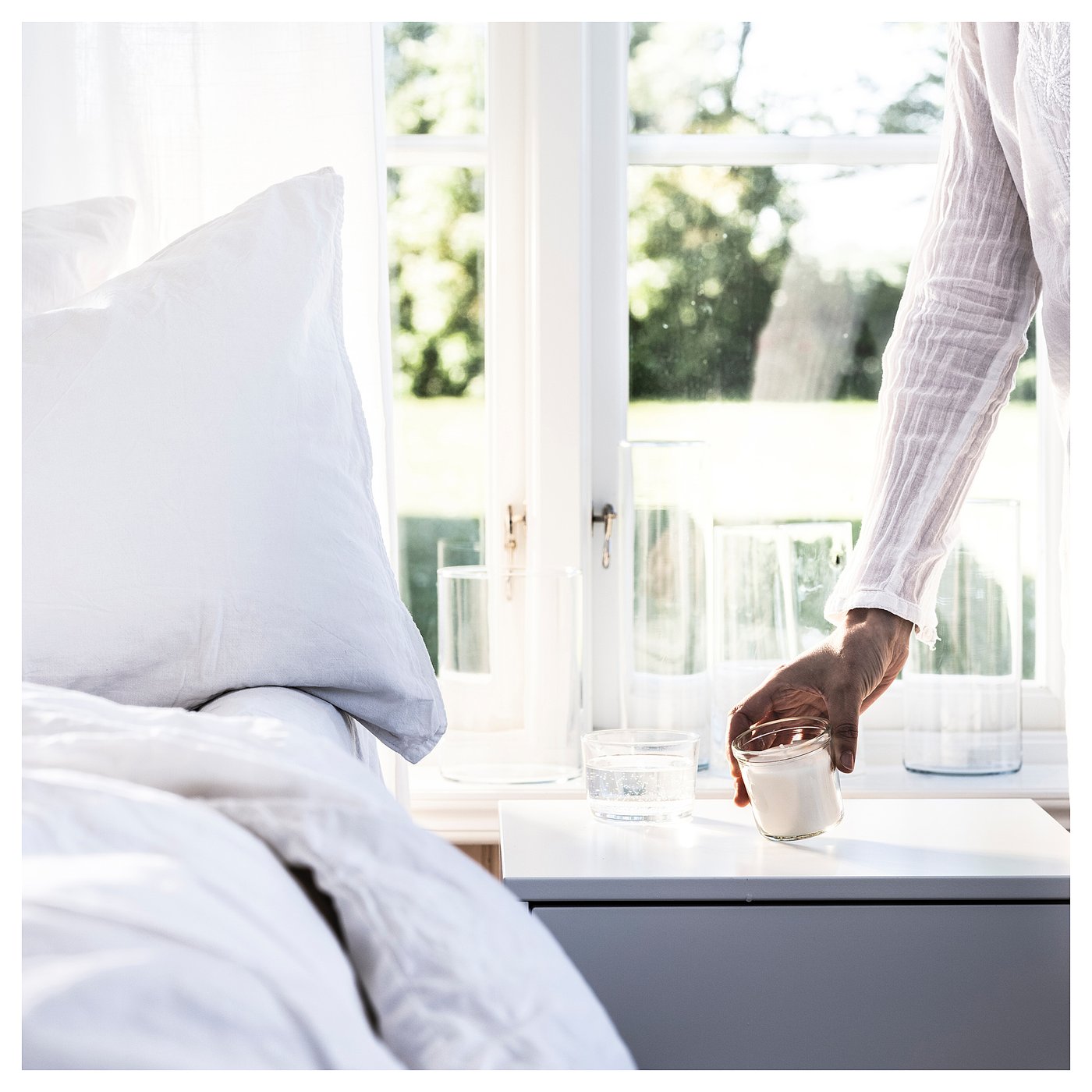 A person holds a glass near a window beside a white pillow. Sunlight streams in, illuminating the scene.
