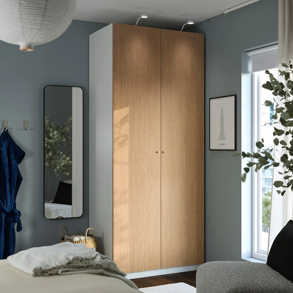Cosy bedroom with light wooden PAX wardrobe, blue wall, grey mirror, and vibrant art. Comfortable with white rug and grey armchair.