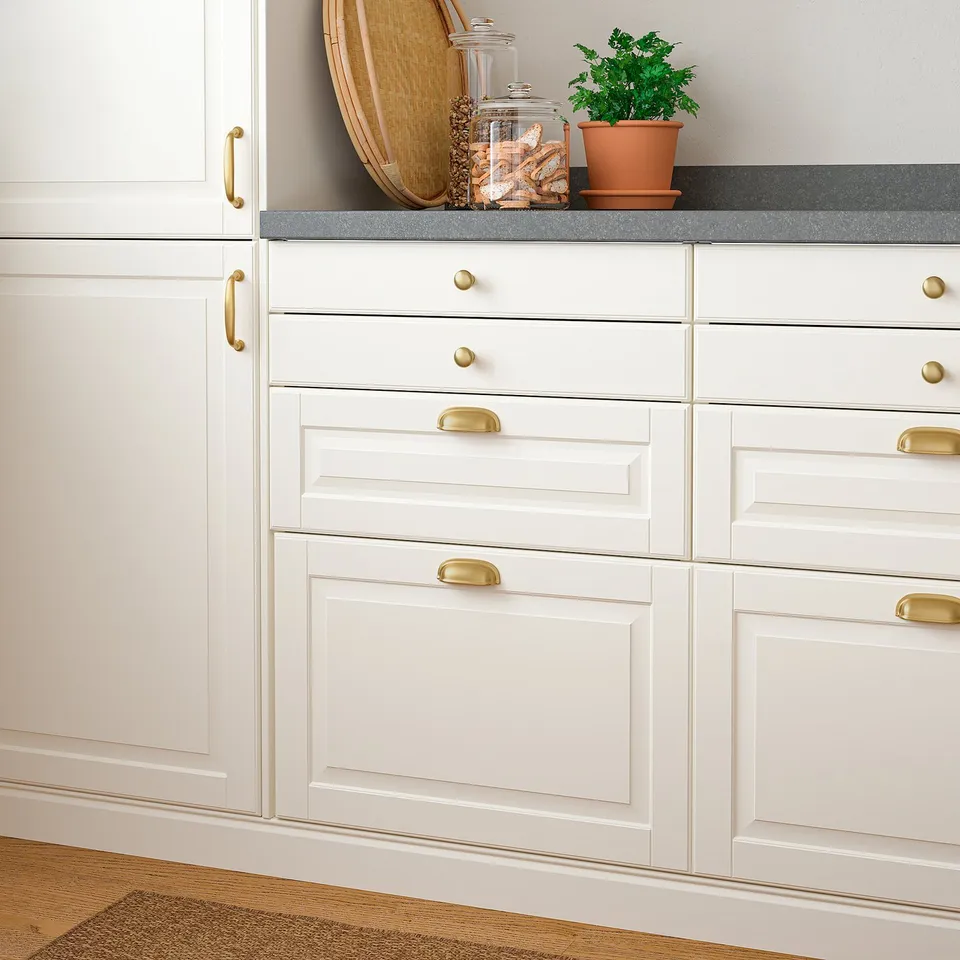 White kitchen cabinets with gold handles, a pot with green plant, and a jar filled with breadsticks on a grey countertop.