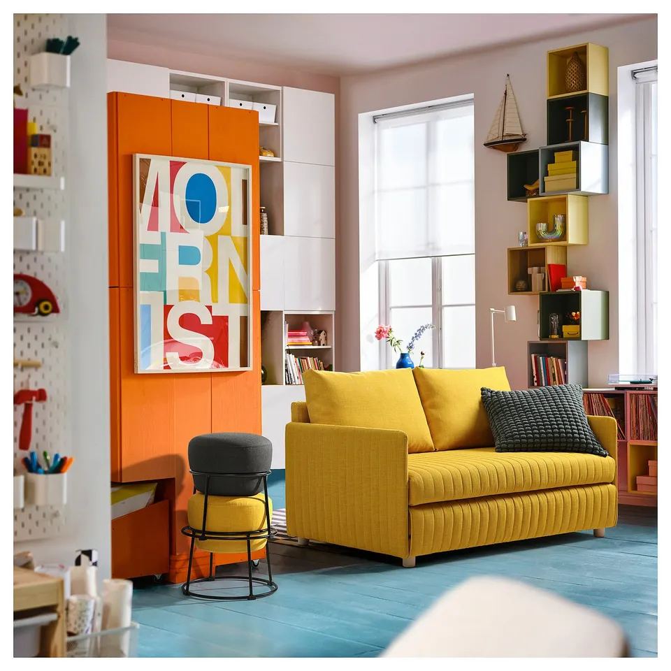 Bright, modern living room with yellow couch, colourful art, and round, grey footstool.
