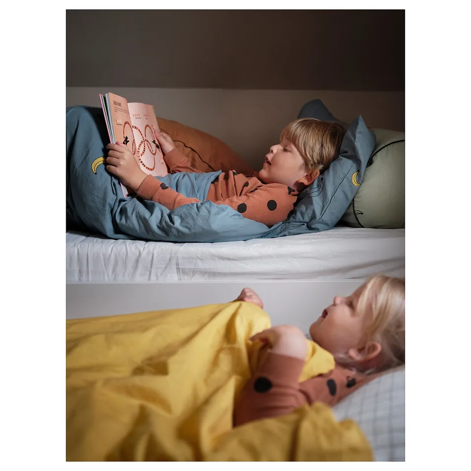 Two kids nap in blue and yellow SLÄKT bunk beds, top child reading a book.