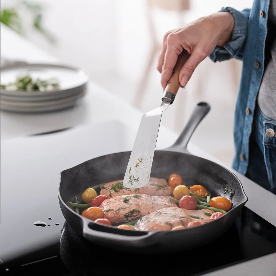 Person cooking fish with VARDAGEN spatula in skillet, surrounded by tomatoes and herbs.